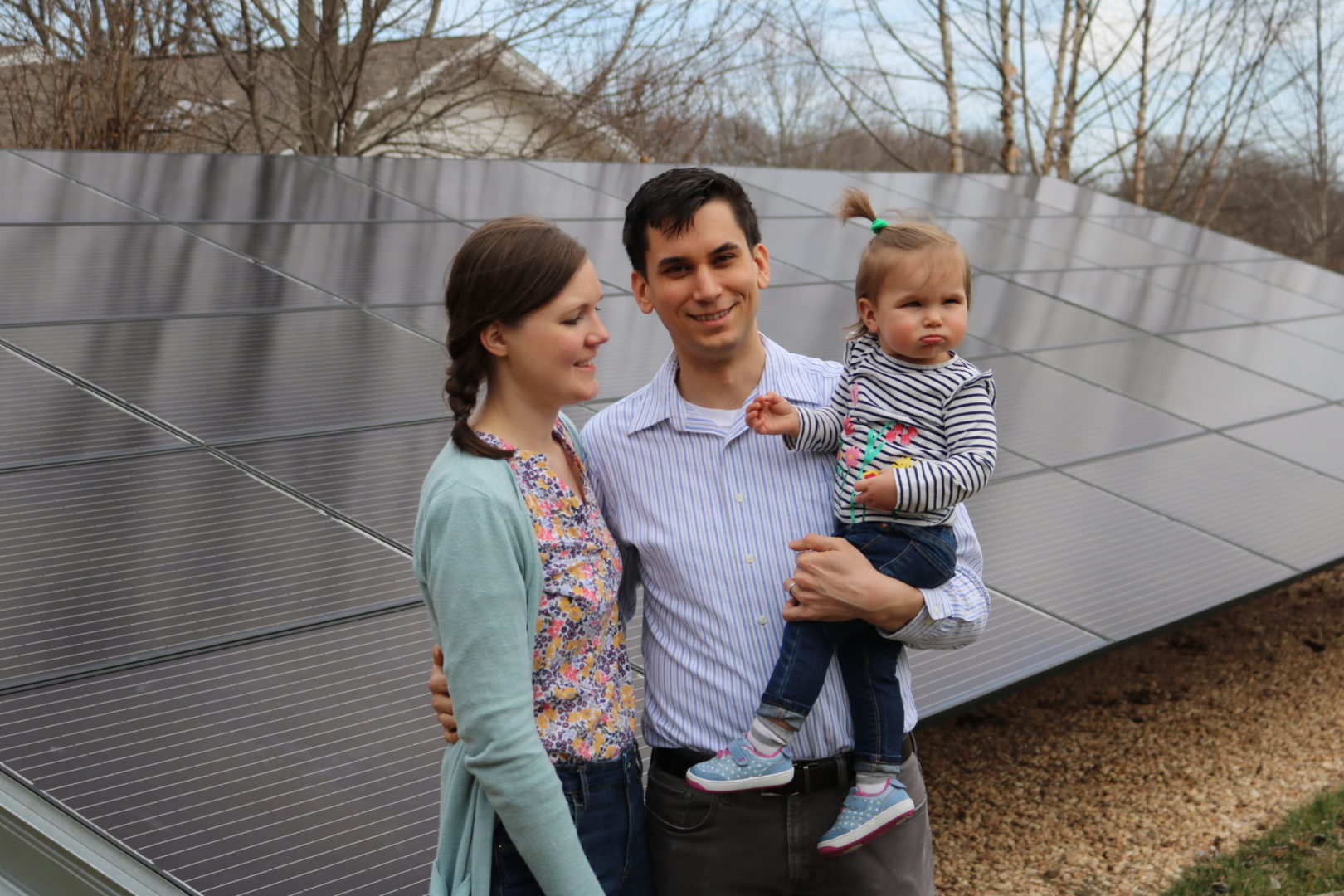 A young couple holding their baby while standing in front of a solar panel ground mount