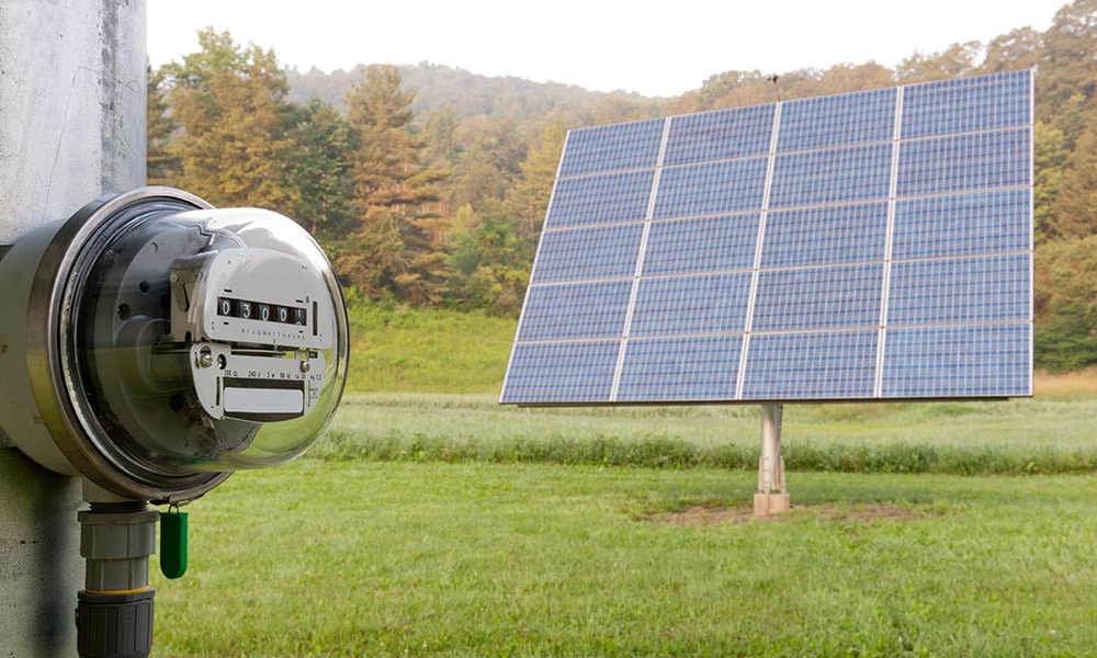 An electric meter is in focus. A ground-mounted solar panel stands in the background.
